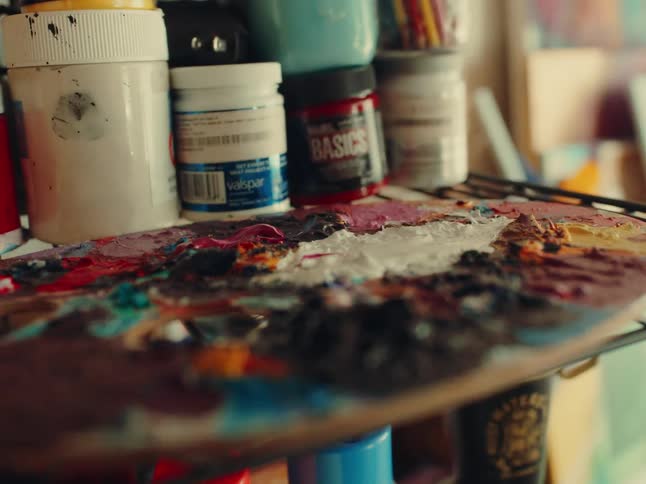The video shows a closeup of an artists palette with thick textured paint on it the palette is surrounded by jars and bottles of paint and art supplies