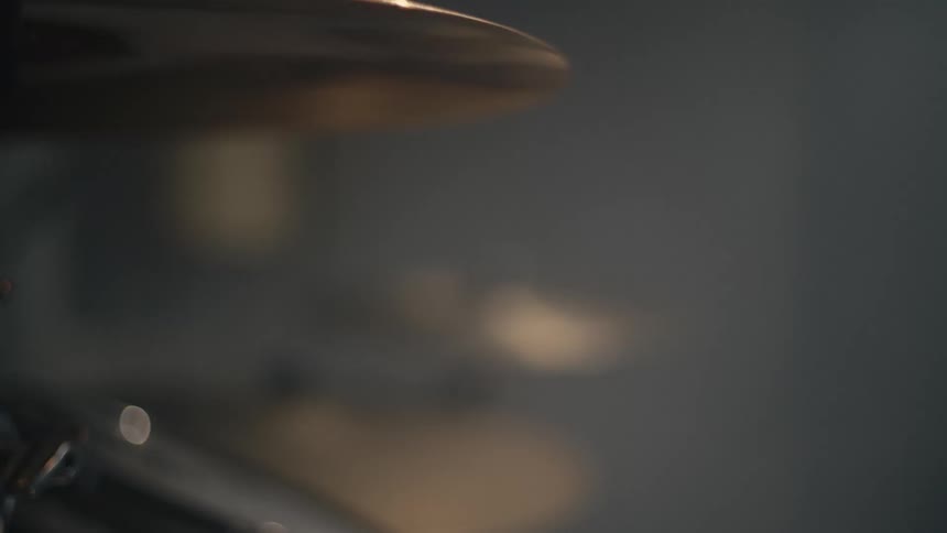 A closeup shot of a drummers hand playing a cymbal with a drumstick the cymbal vibrates slightly after being struck and other parts of the drum kit are visible in the blurry background