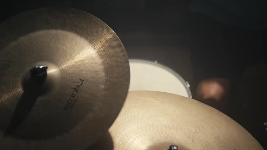 A drummer wearing a black wristband strikes a large golden cymbal with a drumstick in a dimly lit room