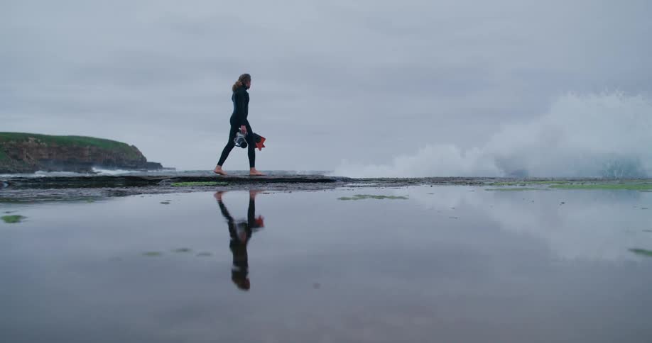 Person in a wetsuit walks barefoot on the beach holding a surfboard with their reflection visible in the shallow water