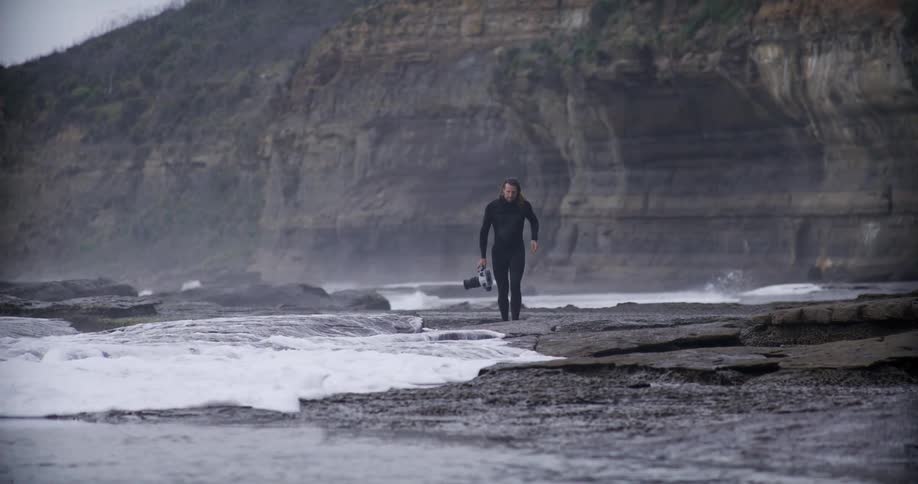 Man in swimming suit walking on rocky shore with professional video camera