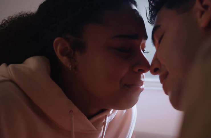 Teenage boy wipes the tears of a teenage girl before kissing her while they sit near a window during the daytime