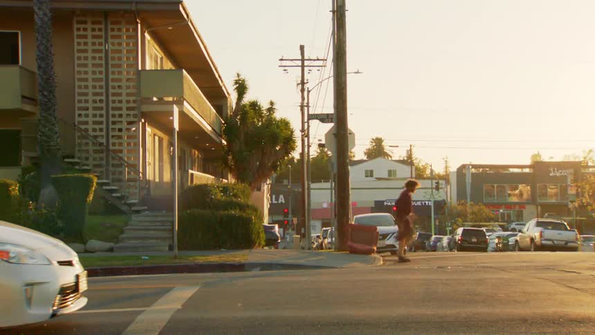 A street corner basks in goldenhour light with light traffic and a person walking withpurpose past a midcentury style apartment building