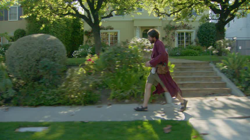 Man in a burgundy robe and sandals walks on the sidewalk in front of a yellow house with a garden