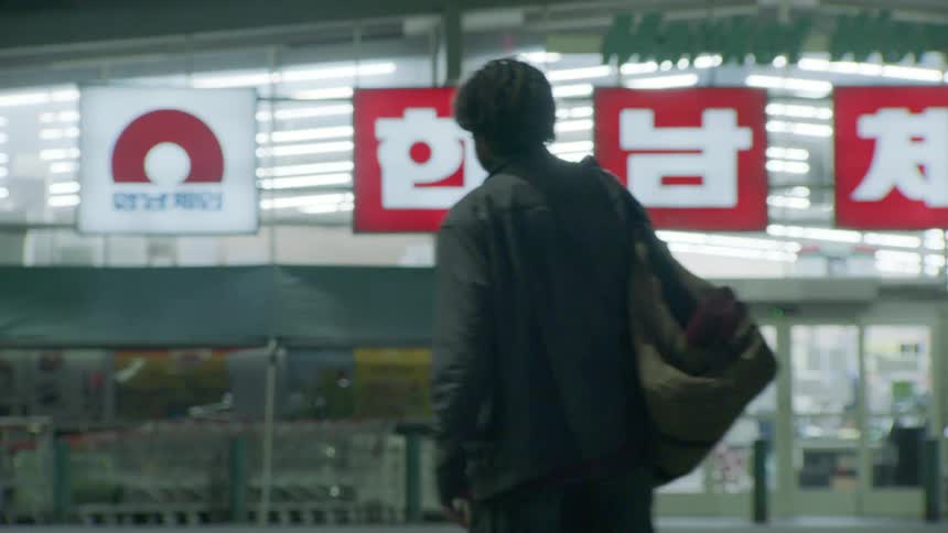 Person carrying a shoulder bag walks toward a brightly lit market with red and white signs in korean characters