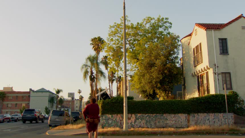 A man wearing a red bathrobe and a utility belt crosses the street in front of a house with a red tile roof in the morning light