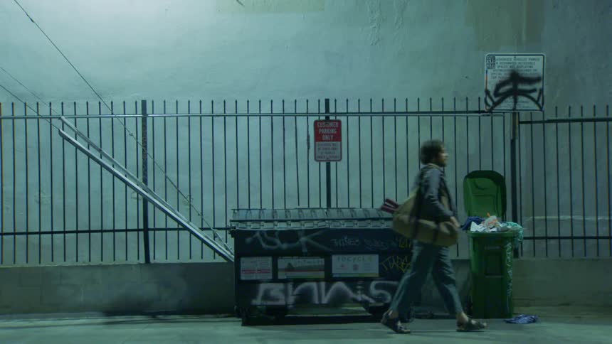 A person walks past a graffitied dumpster and overflowing trash can in a dimly lit alley