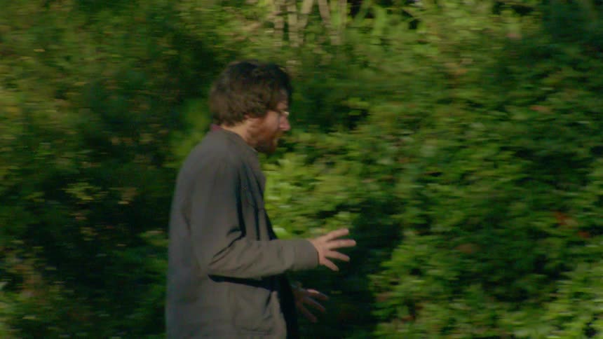 A man with dark hair and glasses walks past a green hedge gesturing as he talks to himself