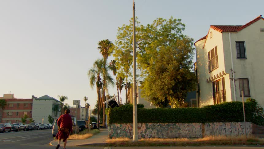 A man running in the middle of the road wearing pajamas suddenly turns back to the sidewalk in the city at sunrise as cars go by