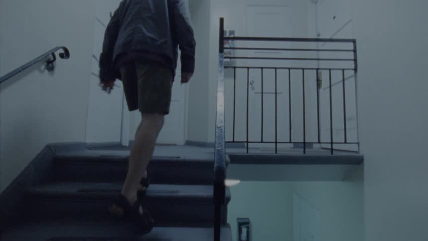 A man wearing a jacket walks slowly up the stairs toward an apartment door
