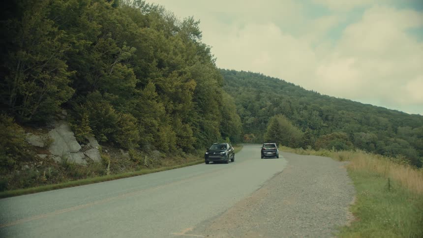 Cars drive along a winding road through a lush green mountain landscape under a partly cloudy sky