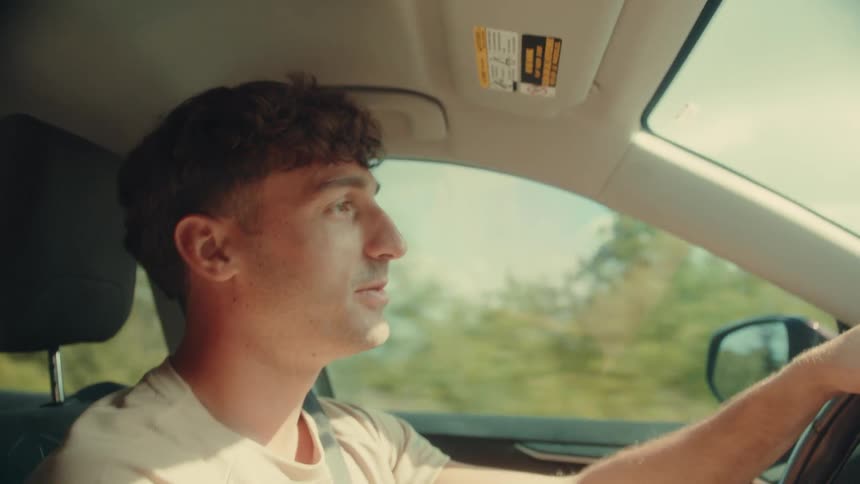 A short video clip shows a man driving a car the video starts with the man looking forward then he turns to look directly at the camera then turns his head back to the front he has brown hair and is wearing a lightcolored tshirt the sun visor is vi