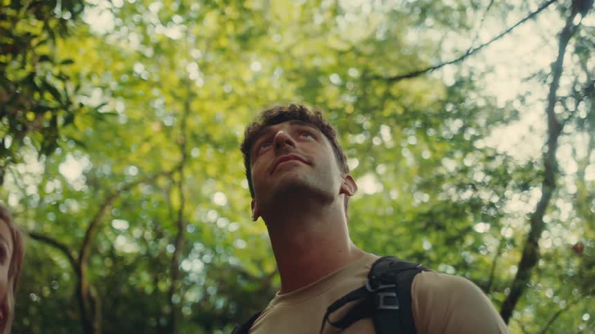 The video shows two people a man and a woman standing outside in a forest one line is the mans gaze which is looking up into the canopy of the trees the second line is a womans jawline which is visible on the left side of the frame as she looks to