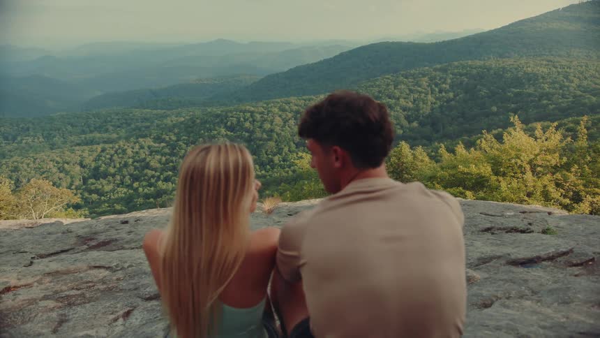 A young couple sitting on a rocky cliff looking out into the wilderness covered in green vegetation at midday
