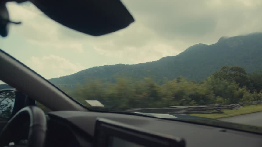 A car cruises along a scenic mountain road offering a view of lush greenery and rolling hills beneath a cloudy sky
