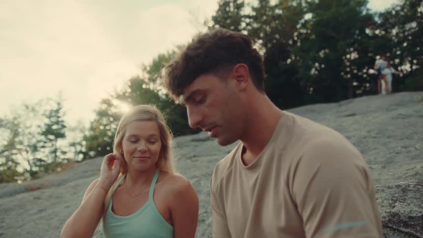 A man and woman are sitting on a rocky outdoor surface seemingly on a break from an activity the woman who has blonde hair and is wearing a light blue tank top is holding a small red item while looking at the man the man with dark hair is looking