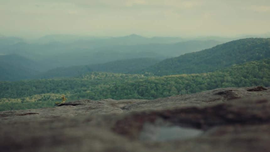 The video shows a wideangle shot of a mountainous landscape with a hazy foggy look the camera is placed on a rocky ground in the foreground with a puddle of water visible the scene slowly pans up to reveal more of the rolling hills and mountains in t