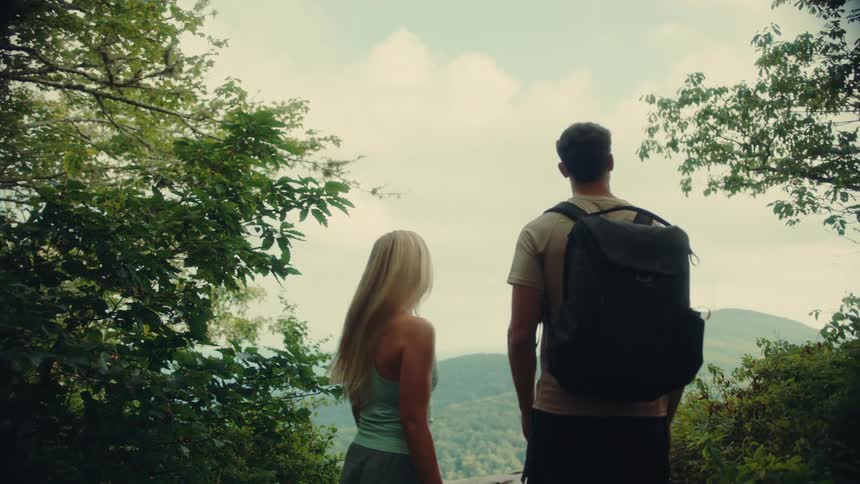 The video shows a couple hiking to a scenic viewpoint overlooking a valley of green rolling hills they stop at the edge of a rocky outcrop framed by trees and admire the expansive view