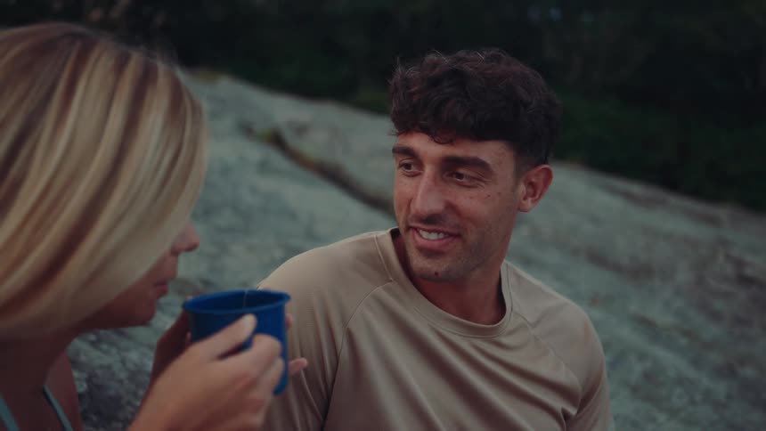 The video shows a man and a woman sitting on the ground likely in a park or forest sharing a drink from a blue metal mug the woman is holding the mug while the man smiles and talks to her the background is a mix of green foliage and a rocky surface