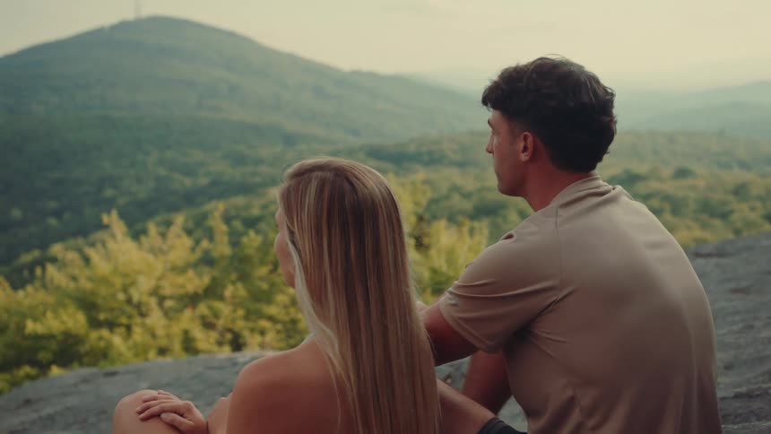 Two friends are sitting near the edge of a rocky cliff admiring the valley below and the forested mountains on the horizon
