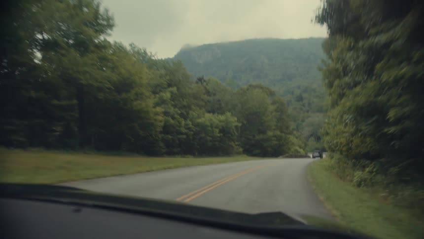 The video shows a firstperson perspective from inside a car driving on a winding twolane road through a rural area the road is surrounded on both sides by dense green trees and in the distance a mountain range is visible under a cloudy sky