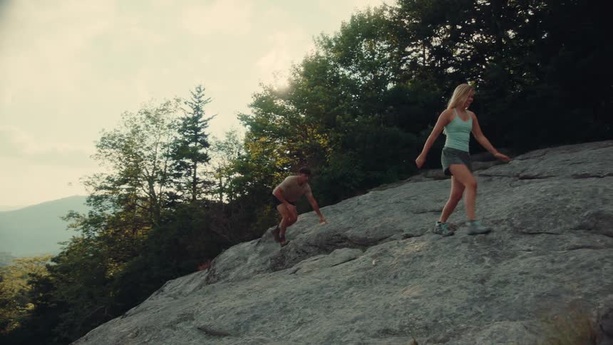 A young man and woman climb a rocky mountain in a forest area during daytime