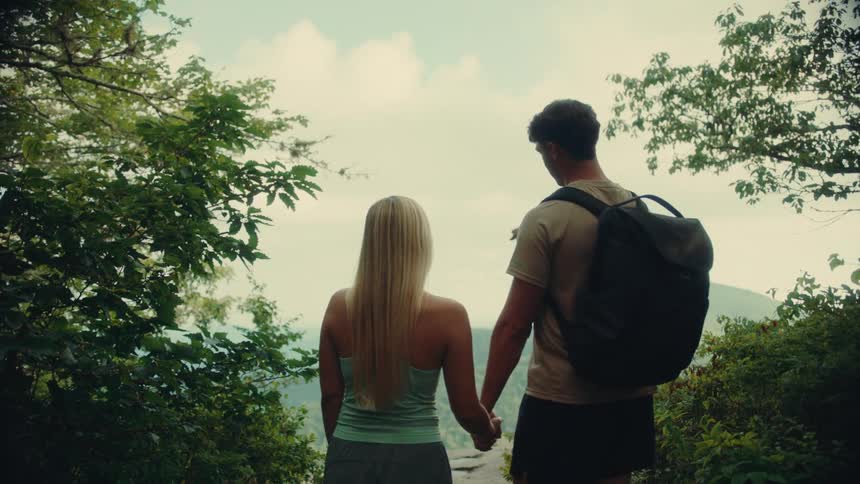 A couple holding hands stands at a scenic overlook surrounded by lush greenery gazing at distant mountains on a cloudy afternoon
