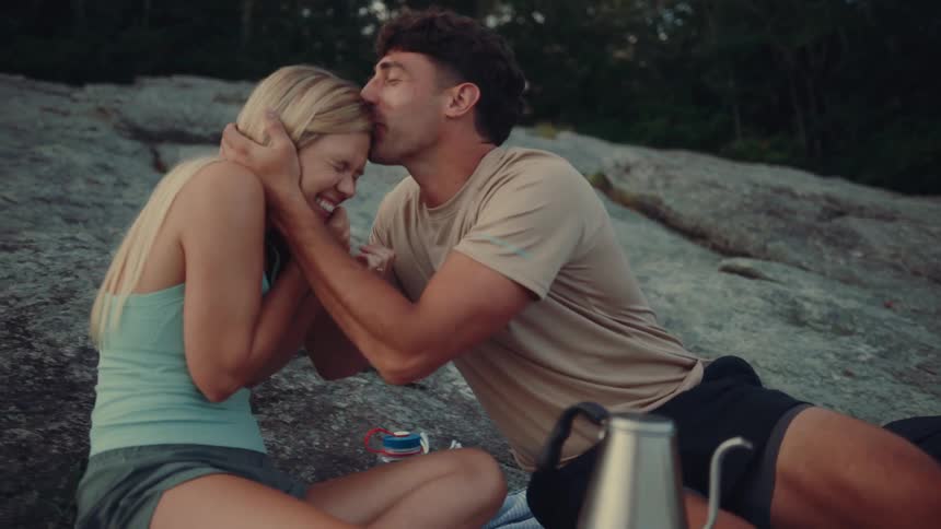 A short video clip shows a man and a woman sitting on a rock playing a hand game and laughing the man and woman then lean in and kiss they are both smiling and appear to be enjoying each others company