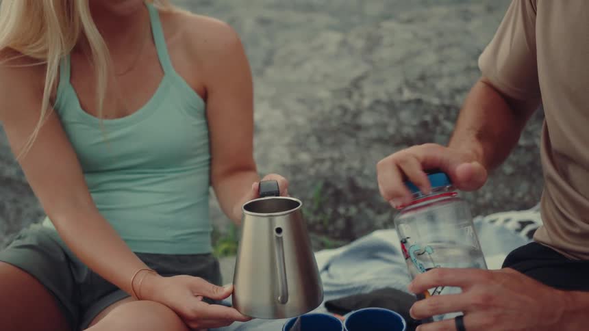 A man pours water from a glass jar into a metal kettle while a woman steadies it during a relaxed outdoor picnic