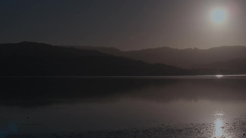 A lake surrounded by hills and mountains with the orange sun setting in the background