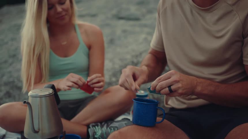 The video shows a man and a woman sitting on the ground likely in a park or forest sharing a drink from a blue metal mug the woman is holding the mug while the man smiles and talks to her the background is a mix of green foliage and a rocky surface