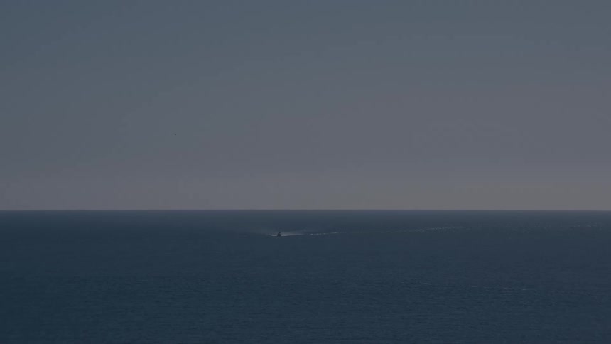 A distant boat leaves a faint trail across the calm ocean under a hazy afternoon sky