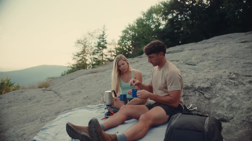 A man and woman are sitting on a rocky surface possibly on a mountaintop enjoying a hot beverage the man is pouring something from a metal pot into a blue mug while the woman sips from her own blue mug they are both looking at each other and smiling