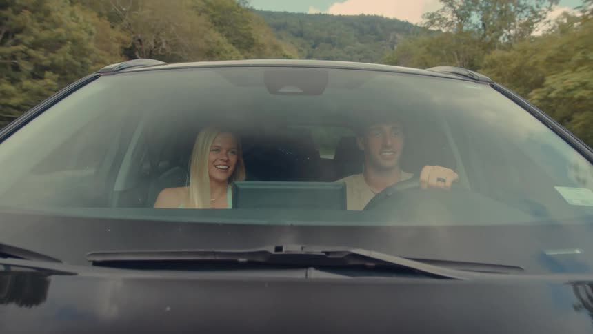 Man and a woman inside a car traveling along a road surrounded by forested mountains