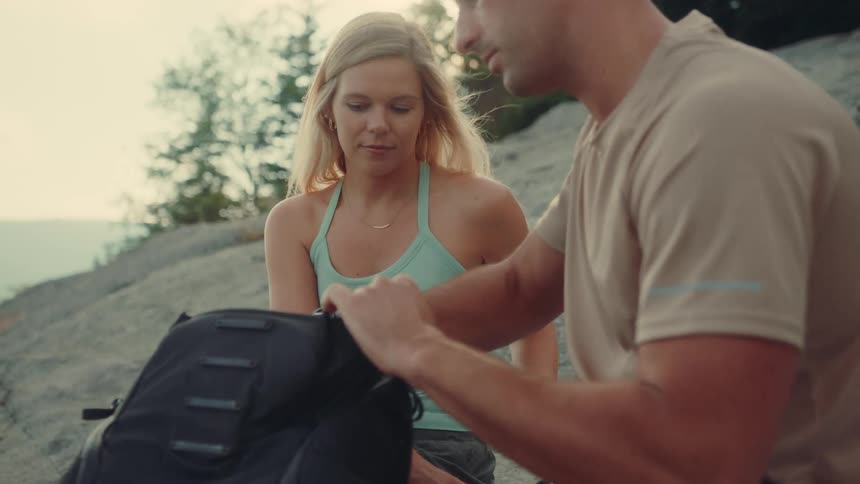 Man and a woman sit on a rock outdoors taking mugs and hiking gear out of their backpacks