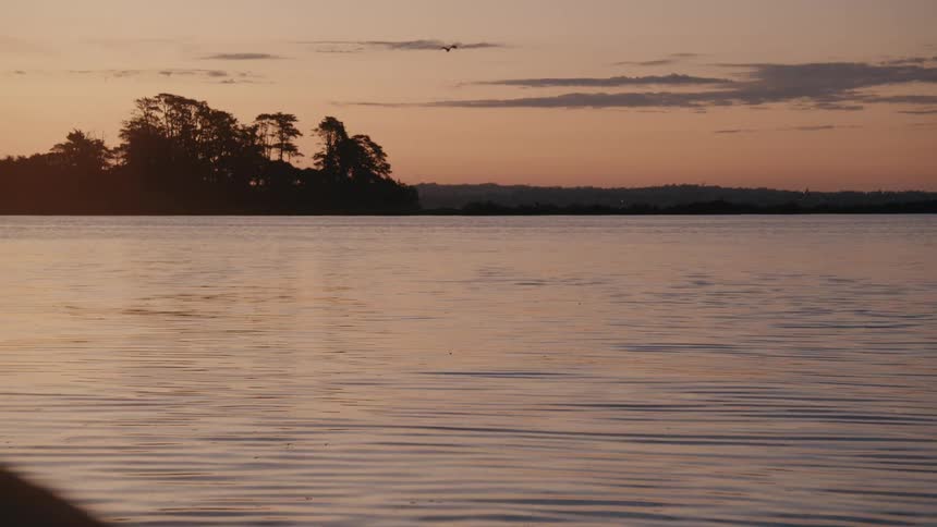A tranquil body of water reflects the warm hues of sunset as trees silhouette an island in the distance