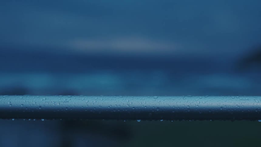 Raindrops cling to a metal railing in focus against a blurred moody blue seascape