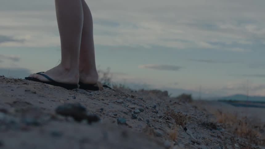Bare legs in flipflops stand on a rocky dry landscape under a cloudy sky