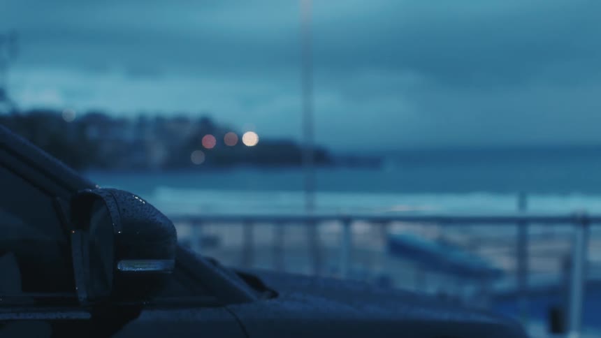 The video shows the side mirror and fender of a parked car with the ocean and a blurry shoreline in the background the scene is dark and appears to be shot at dusk with a cool blue color grade