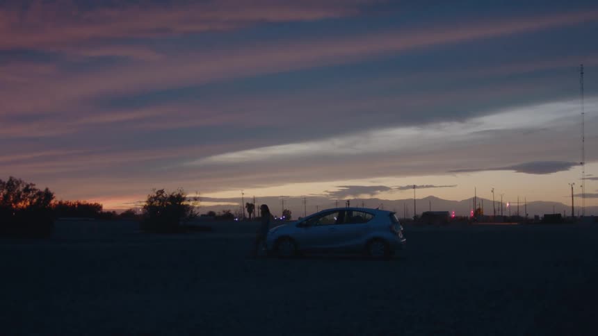A lone car sits parked in an open lot under a dramatic dusk sky streaked with purples and oranges