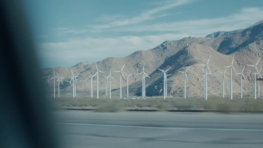 A long row of white wind turbines stands in front of barren rocky hills as the view from a moving car passes a highway and a billboard