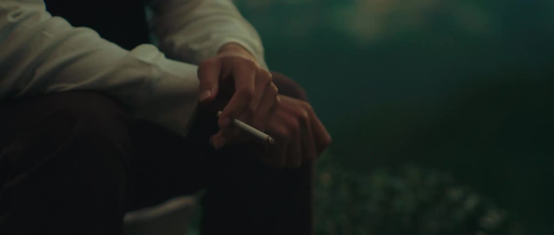 A man in a suit jacket and white shirt holds a cigarette in his hands with the focus on his hands the background is dark and out of focus he is not seen smoking the cigarette but simply holding it between his fingers