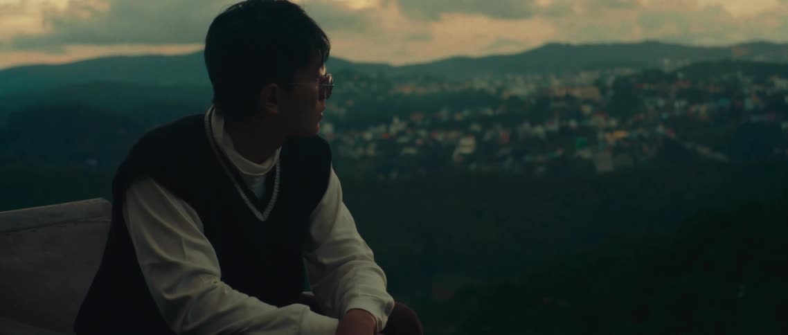 A short video clip shows a person sitting outdoors looking out at a hilly landscape with a city in the distance the person is wearing glasses and a sweater vest over a white longsleeve shirt they are smoking a cigarette and seem to be deep in thought
