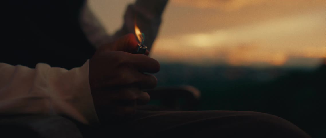 The video shows a closeup of a persons hand holding a lighter which is being flicked on and off the background is a softfocus landscape with an orange and yellow sunset the warm colors of the sunset contrast with the darker silhouette of the person