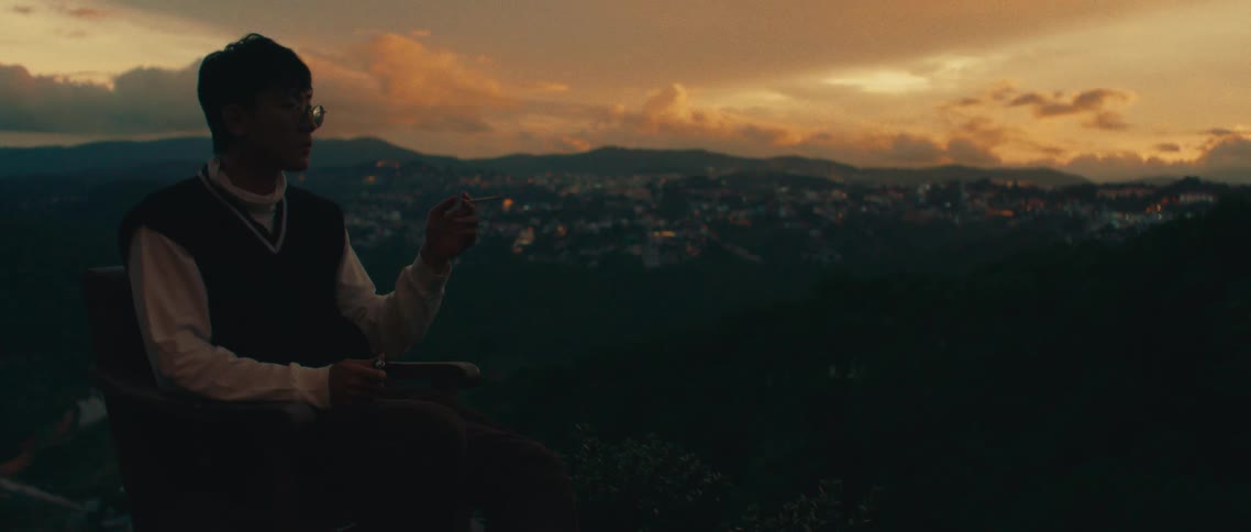 A man stands calmly with a cigarette in hand gazing over a glowing hillside city at sunset