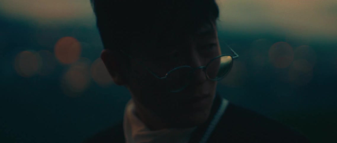 A short video clip shows a person with glasses standing outside at dusk looking out into the distance the background is a city with blurred lights suggesting a lowlight environment the video is a closeup shot of the persons face from the side