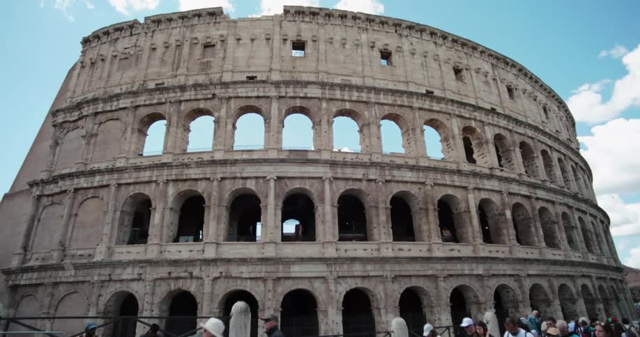 A large crowd of people walking in front of the ancient colosseum a tiered stone structure with arches and a damaged facade all under a clear blue sky