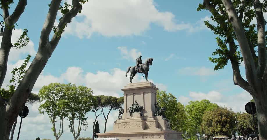 A video clip shows a statue of a person on a horse in a town square framed by trees on either side the statue is on a large ornate stone base with other smaller statues on it the sky is bright and blue with some white clouds