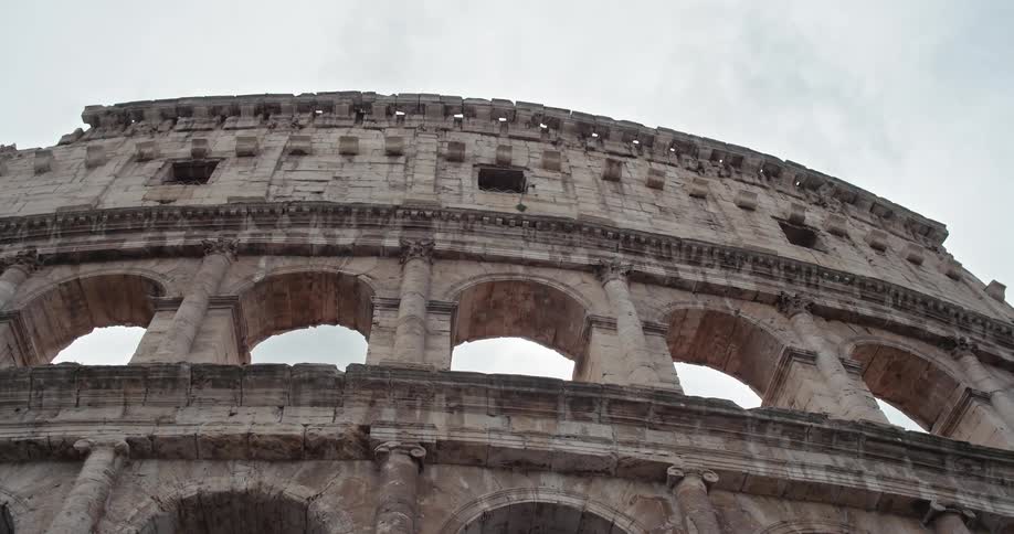 The video shows a lowangle shot of the top section of the colosseum in rome the ancient structure is made of stone and features multiple arches and windows the sky above is overcast and gray which provides a dramatic backdrop for the historical buildi