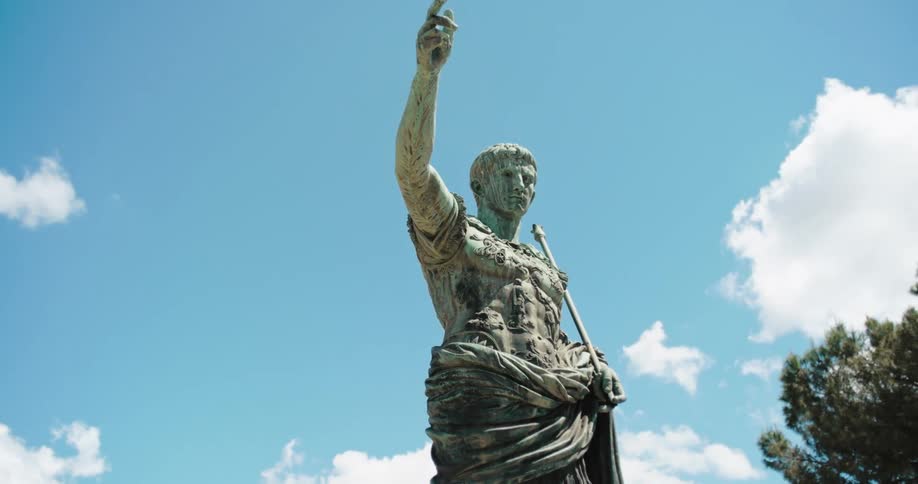 A large bronze colored statue of a man in roman armor holding a staff stands under a clear blue sky